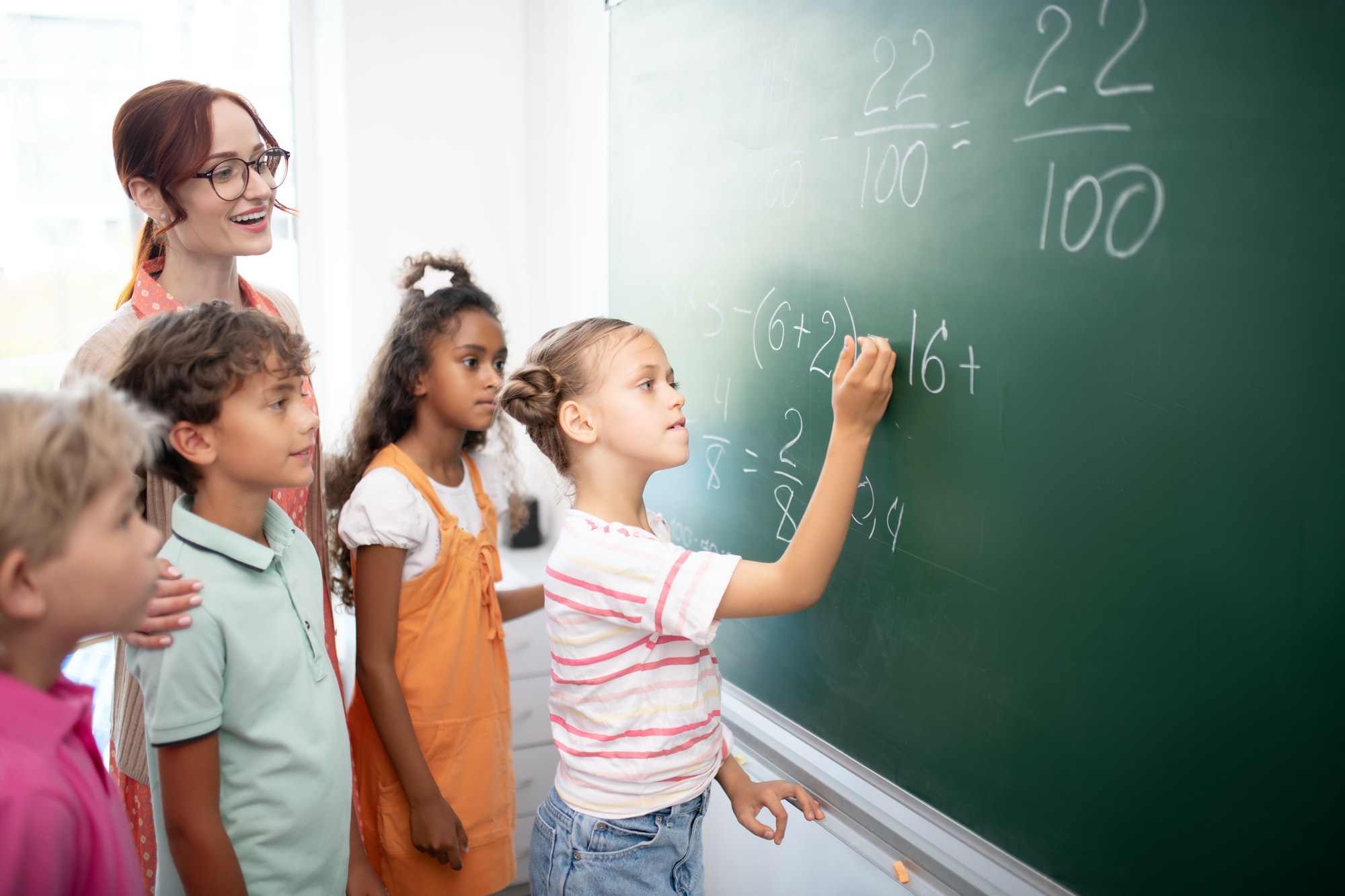 Smart girl writing down numbers on blackboard during math class